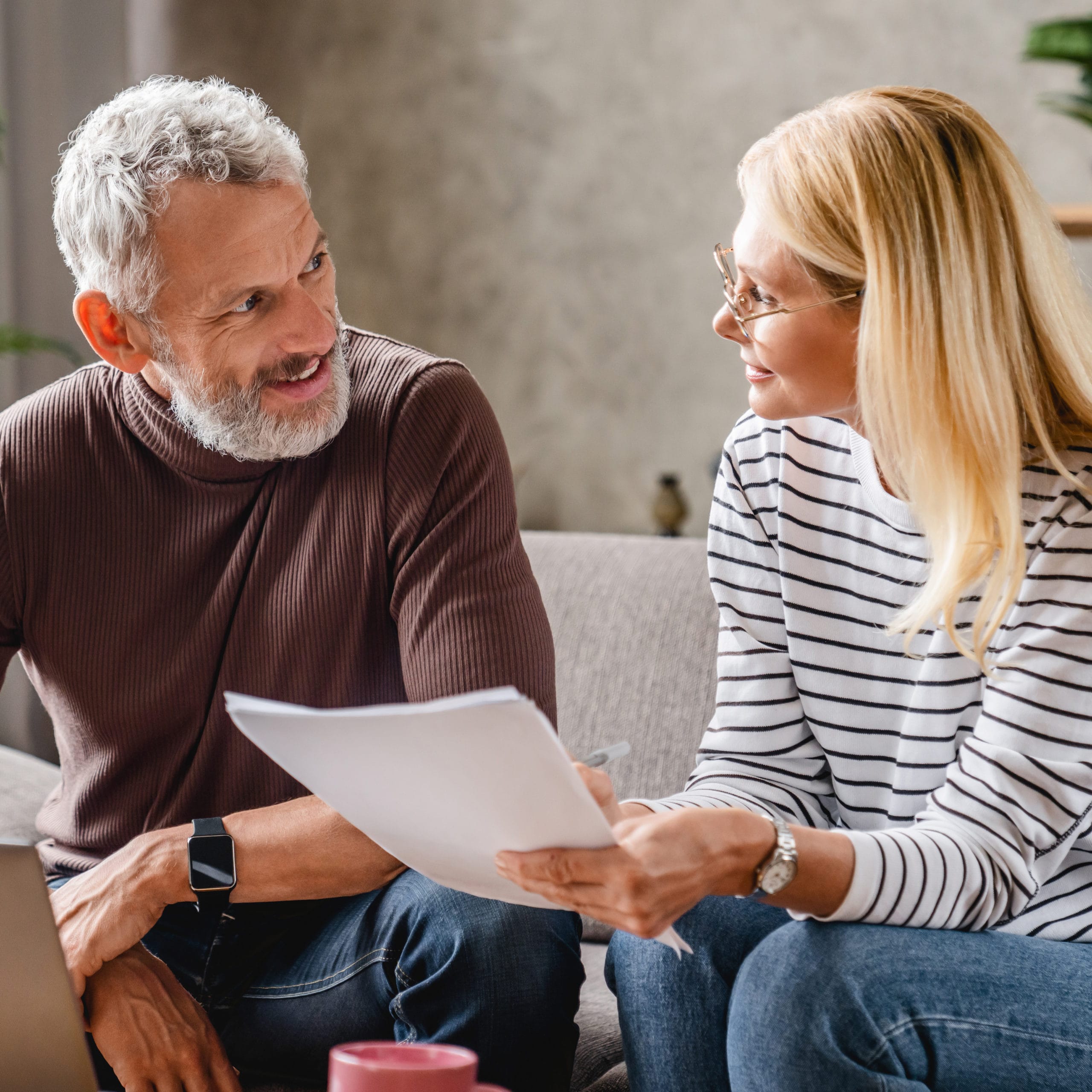 Smiling middle aged couple with laptop studying documents while working on couch at home